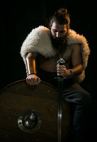 A man dressed in a Viking inspired outfit holds a Norse sword, wearing a fur cloak and looking towards the camera