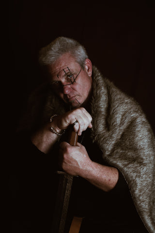 A man dressed in Viking style clothing stands against a black background facing the camera. He wears a Viking style bracelet and carries a sword.