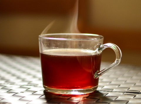 A clear glass mug on a metal table, filled with steaming tea, showcasing the warm amber liquid against a softly lit background, with the steam rising gently above, indicating a freshly brewed hot beverage.