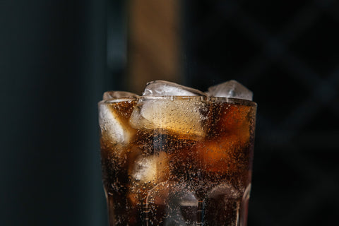 A close-up view of a glass of iced cola, with bubbles clinging to the ice cubes and the side of the glass, highlighting the carbonation and the dark, rich color of the soda against a blurred background.