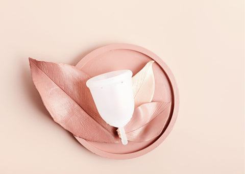 On a pale pink background, a reusable menstrual cup sits on a small circular pink tray