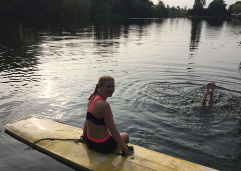 Sarah sits on the edge of a light wooden plank. Around her is the dark blue waters of the lake. You can see a fellow wild swimmers feet just hitting the water in the right corner