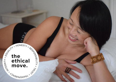 A woman in Y.O.U Underwear's organic cotton black Bralette and boy shorts set lays sideways, smiling, on a bed. In the bottom left corner, the ethical move pledge badge in white sits. 