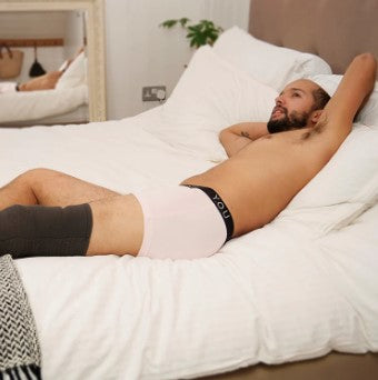 Man lying on a bed wearing a pair of pink hipster trunks. 