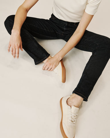 A lady sits on the floor wearing black Vaela jeans, a white t-shirt & trainers