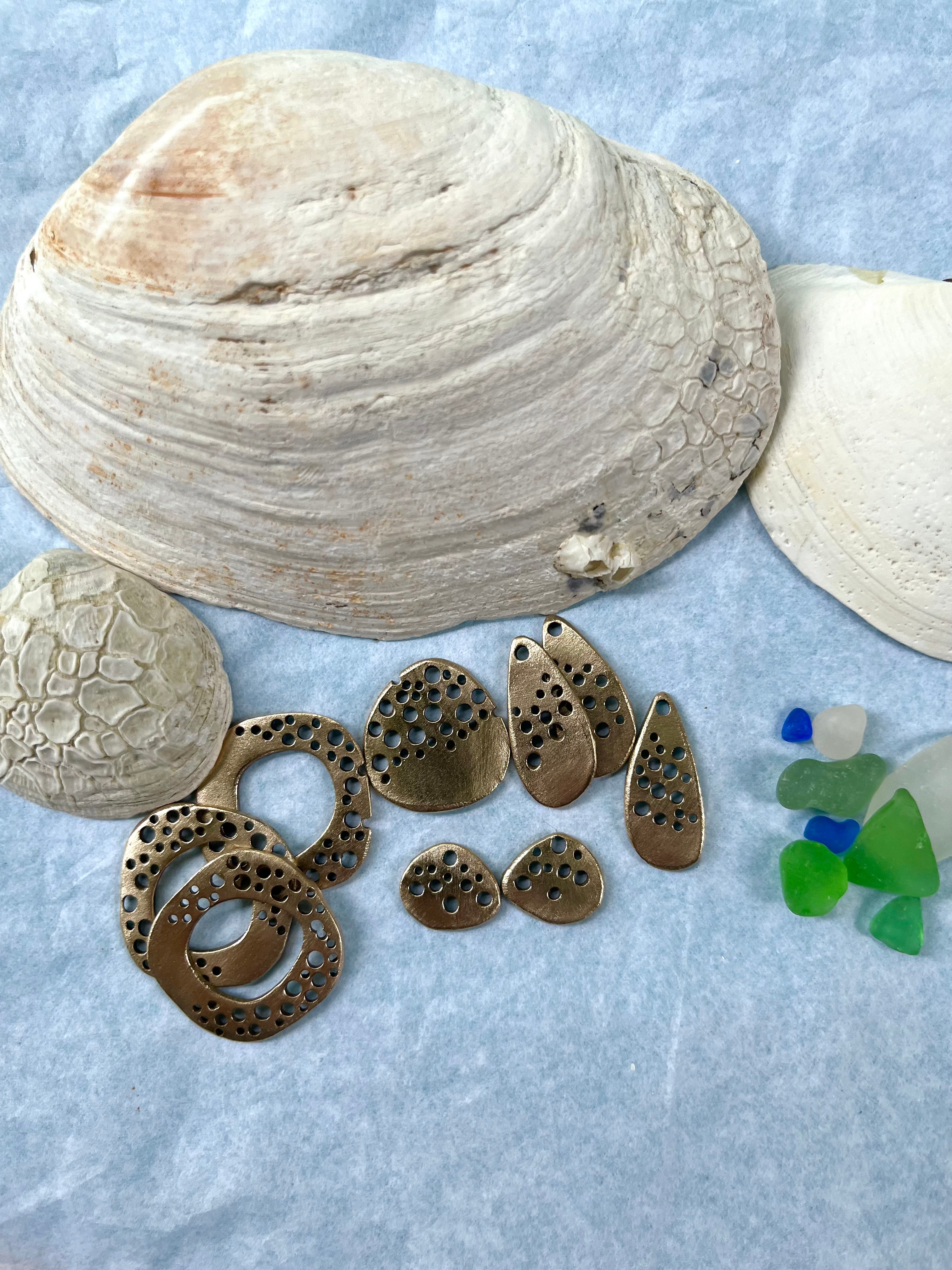 Bronze hand made jewelry charms in front of seashells and beach glass