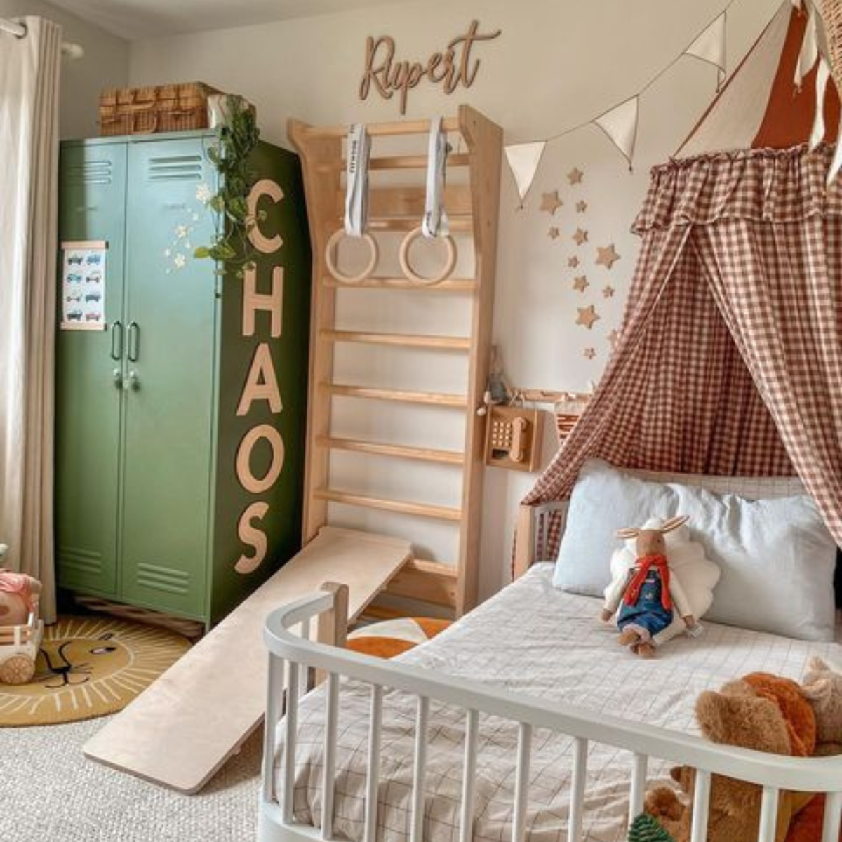 An Olive Twinny has the word CHAOS stuck in large letters to its side. Beside it there is a small wooden climbing gym and a childs bed with a gingham canopy. Trailing plants hang down from the top of the Twinny.