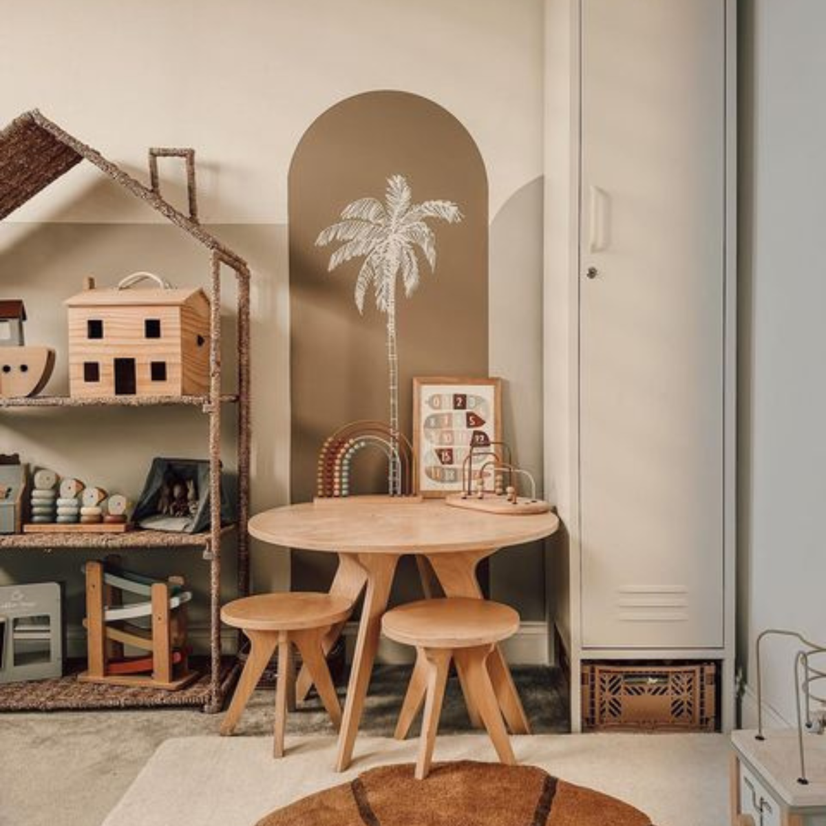 A White Skinny sits in the corner of a neutral-toned room with accents in shades of brown. There is a taupe arch painted on the wall with a white palm tree sketch, and a wooden table and chairs arranged around some toys.
