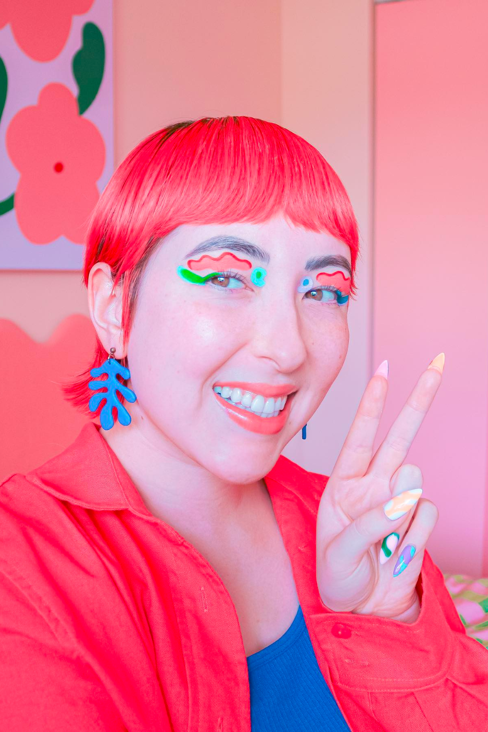 Courn Anh smiles and makes a peace sign at the camera. They have candy pink hair and clothes and wear brightly coloured graphic eyeliner in squiggly shapes with bright blue coral-shaped earrings.