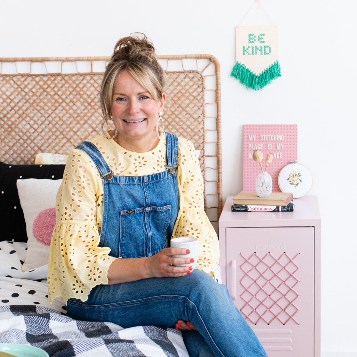 Chloe sits on a bed next to a blush cross-stitched locker. She is wearing denim dungarees with a ruffled yellow shirt and hoop earrings. She is holding a cup of tea.