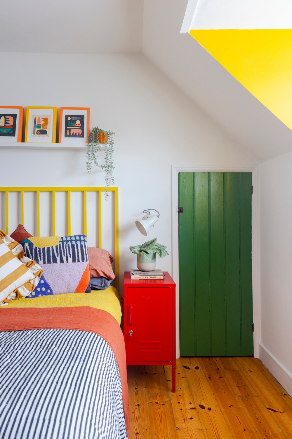 A Poppy red Shorty sits next to a bed dressed in bright, clashing linens. There is a shelf above the bed holding colourful graphic prints and a trailing plant.