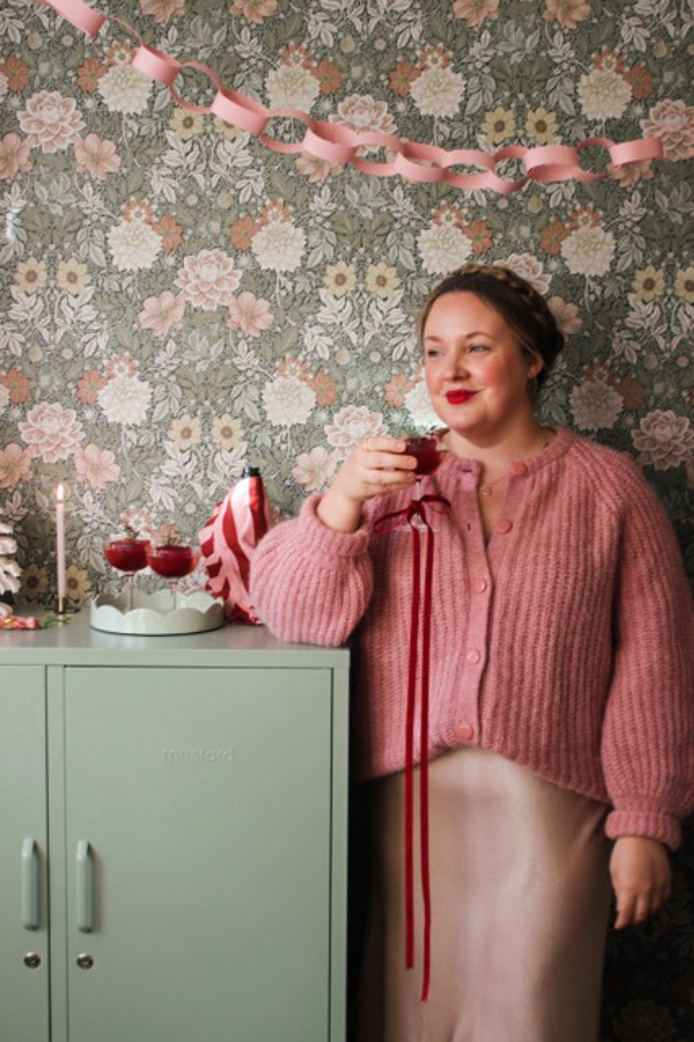 Charlotte Jacklin is wearing a pink cardigan and smiling next to her Sage locker. She is holding a cocktail glass decorated with a velvet ribbon.