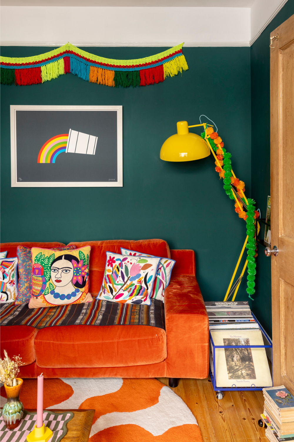A dark teal living room features an orange velvet couch with vibrant colourful cushions. There is a yellow lamp in the corner and a rainbow print on the wall.