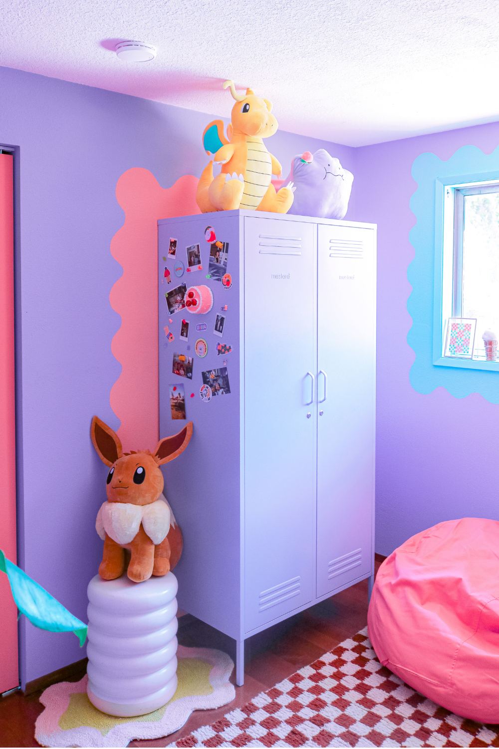 A Lilac Twinny stands in a lilac painted room with a pink wiggly feature painted around it. There are Pokemon stuffed toys next to and on top of the locker, and one side of it is covered in magnets.