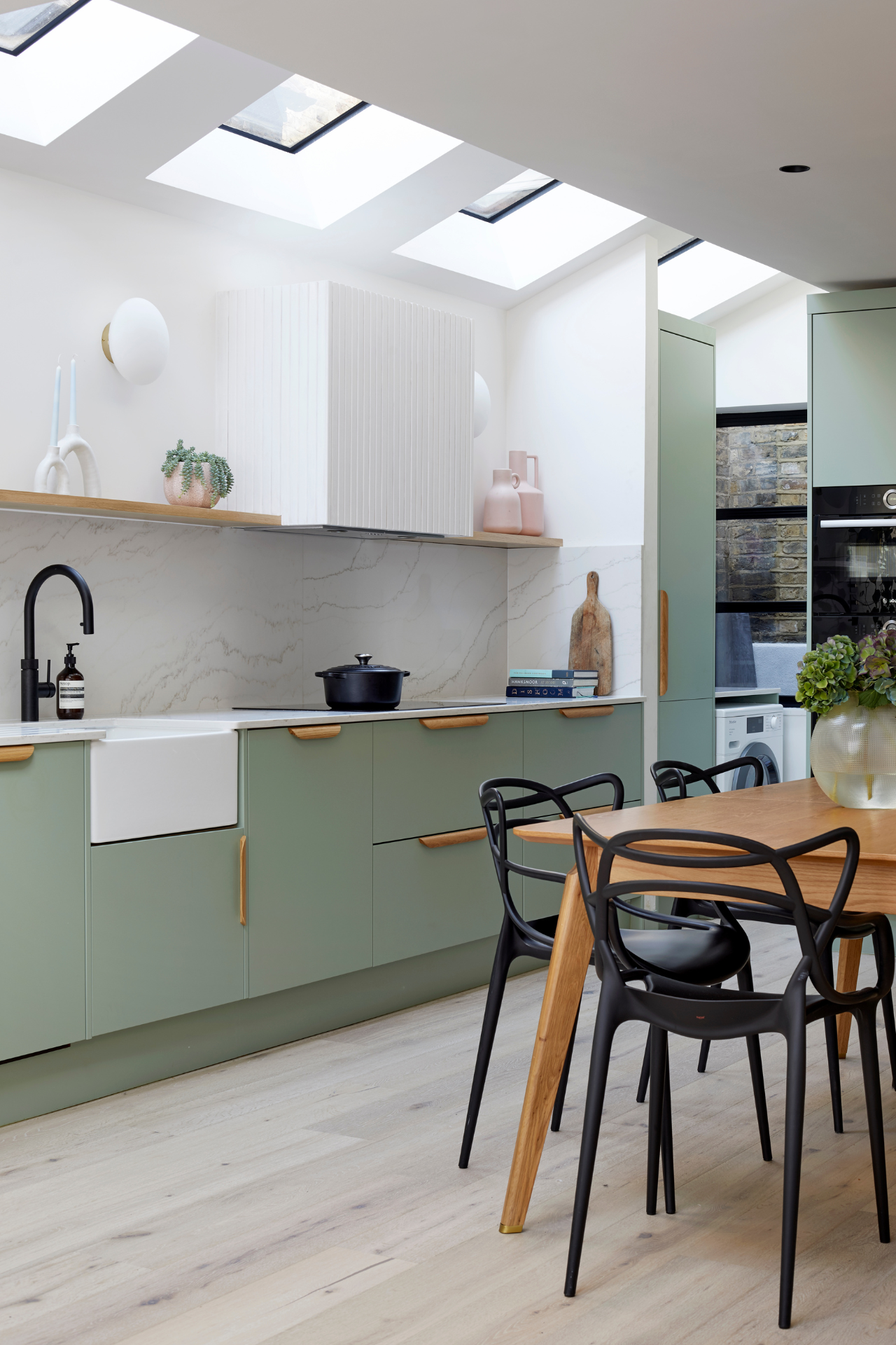 A light-filled kitchen features sage green cabinetry with wood accents. In the ceiling, light filters through a row of skylights.