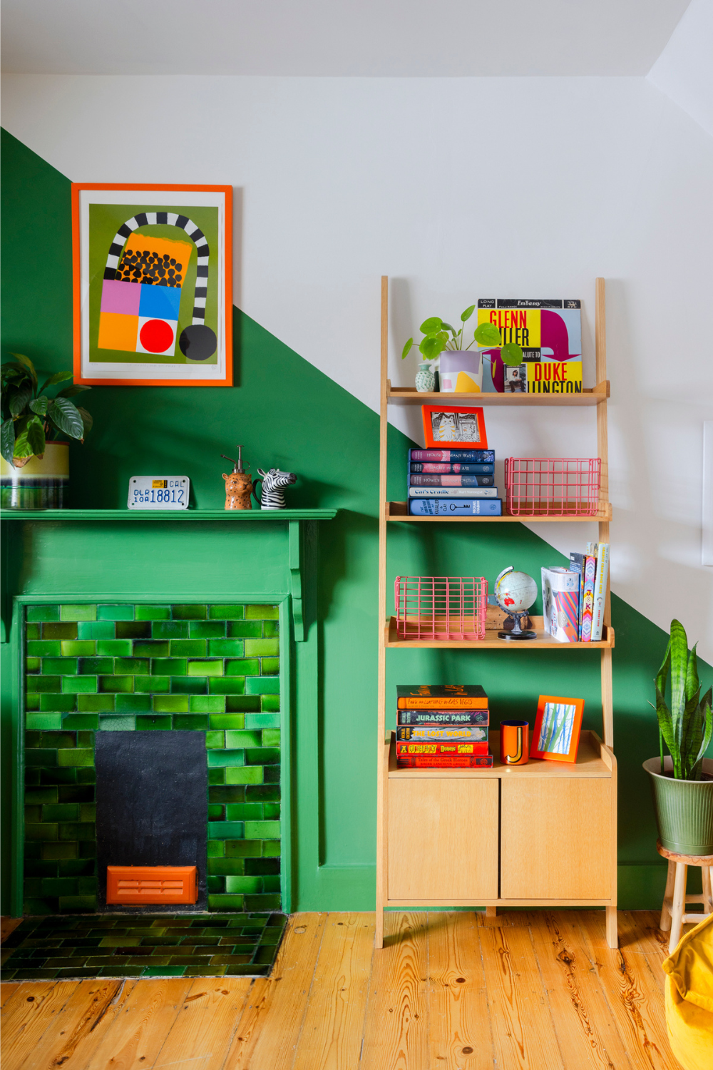 A wooden bookshelf is positioned next to a green tiled fireplace. The wall behind it has a diagonal line down the centre, dividing sections of bright green and white paint.