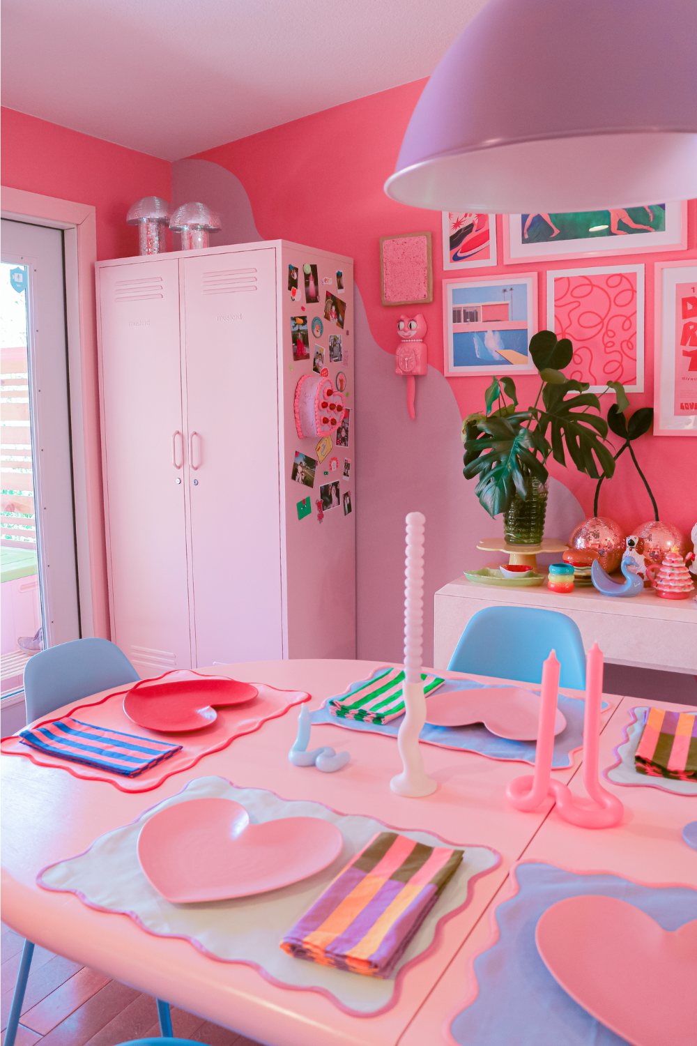 A dining room is painted in blush and candy pinks and a Blush Twinny stands in the corner. It is decorated with magnets and photos and on the dining table there is an assortment of brightly coloured candles and accessories.