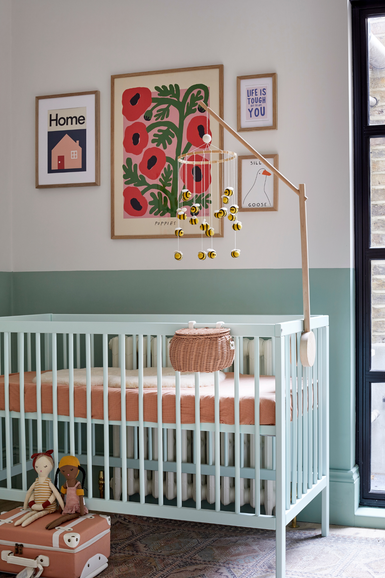 A nursery contains a soft green coloured cot with terracotta sheets. The wall is painted sage green up to halfway, while the top half and ceiling are white. A gallery wall of colourful prints and a bee mobile complete the space.