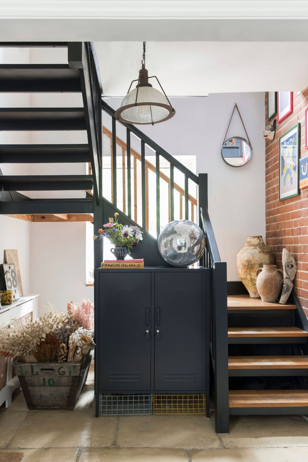 A Midi in Slate sits in a stairwell next to an exposed staircase. On its top there is a large disco ball and a vase of flowers, and next to it a large wooden crate overflowing with dried flowers.