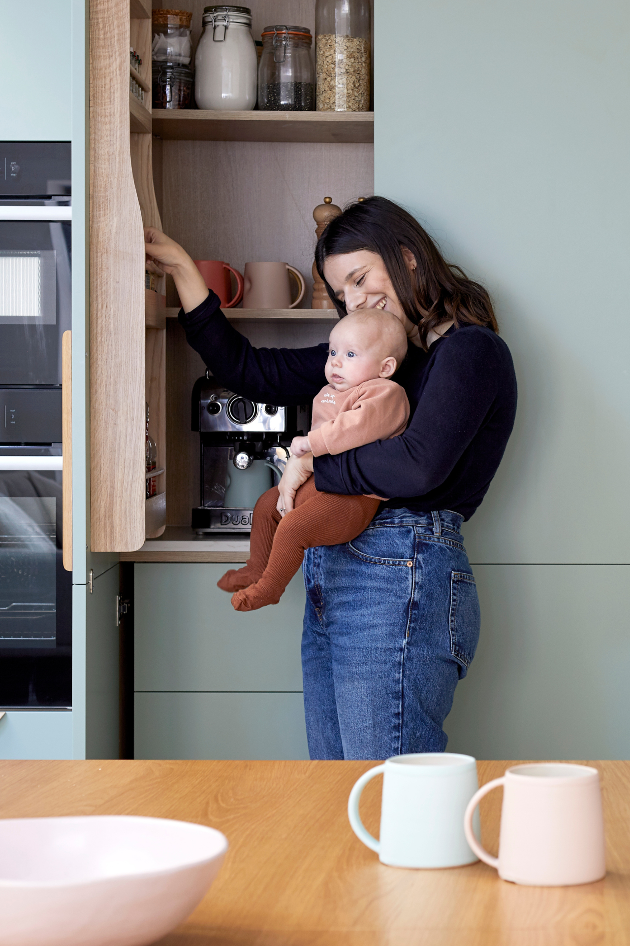 Jess is holding baby Poppy in one arm and smiling as she opens a kitchen cupboard with the other. Inside the cupboard a silver coffee machine sits below a shelf of mugs and pantry supplies.