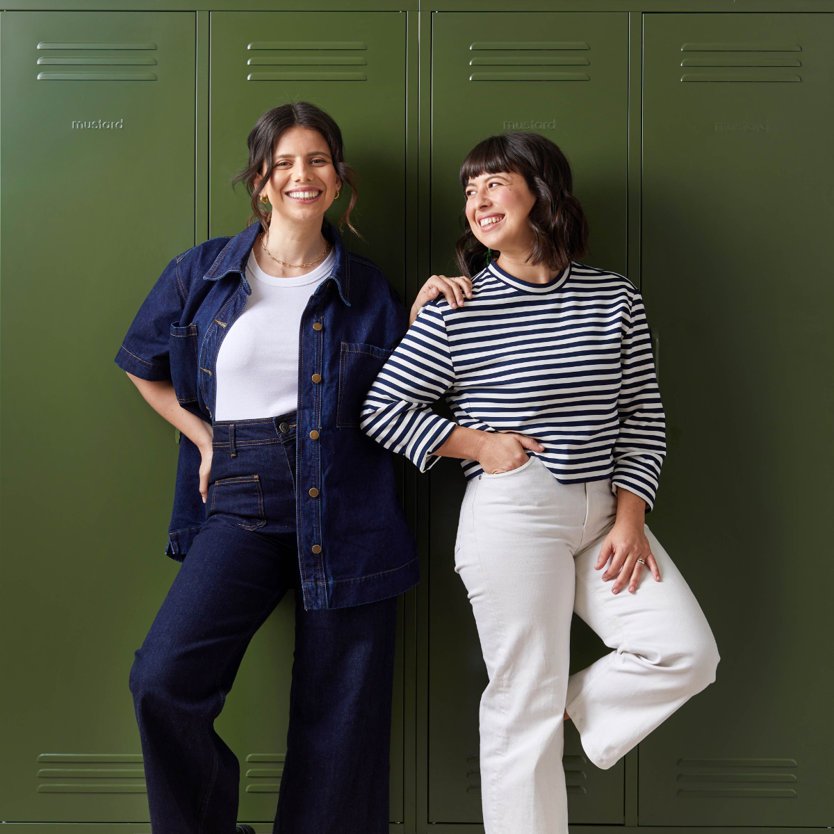 Becca + Jess lean against a wall of Olive lockers. Jess wears a denim two-piece and Becca wears a black and white stripy top with white pants.