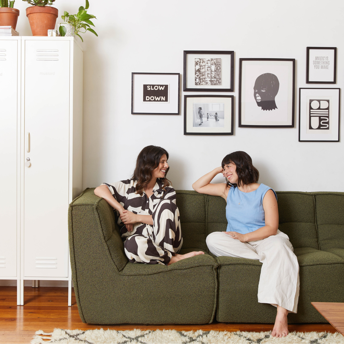 Becca + Jess are smiling and lounging on an Olive couch next to a group of white Skinny lockers. There are black and white artworks on the wall and green leafy plants on top of the lockers.