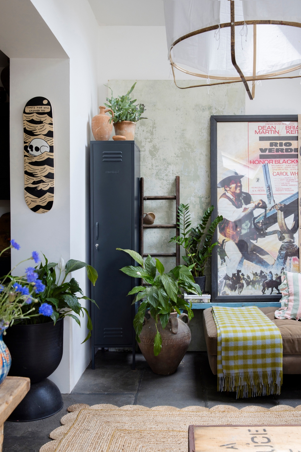 A Slate Skinny is styled in the corner of a plant-filled living space. There is a large vintage movie poster framed against the wall and a hand painted skateboard deck hanging next to it.