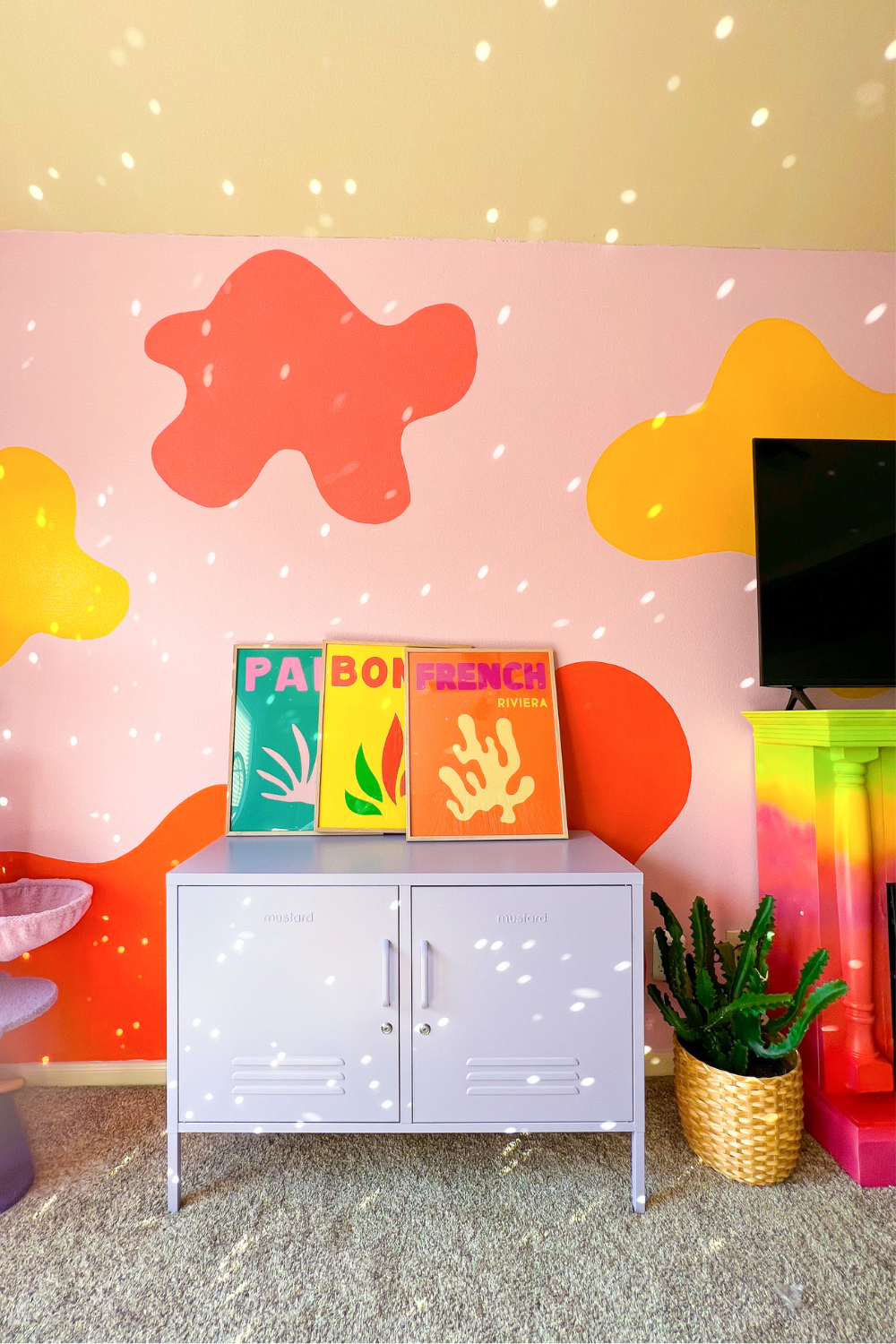 A Lilac Lowdown sits against a pink wall painted with splotches of bright colours. There is scattered light reflecting off disco balls and a trio of Assouline prints resting on top of the locker.