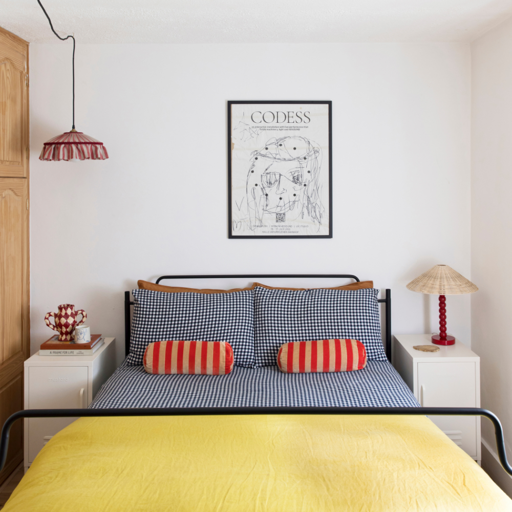 In a white bedroom, a black wrought iron bed is styled in monochrome gingham and bright red and yellow linens. There is a pair of White Shortys nestled either side of the bed.