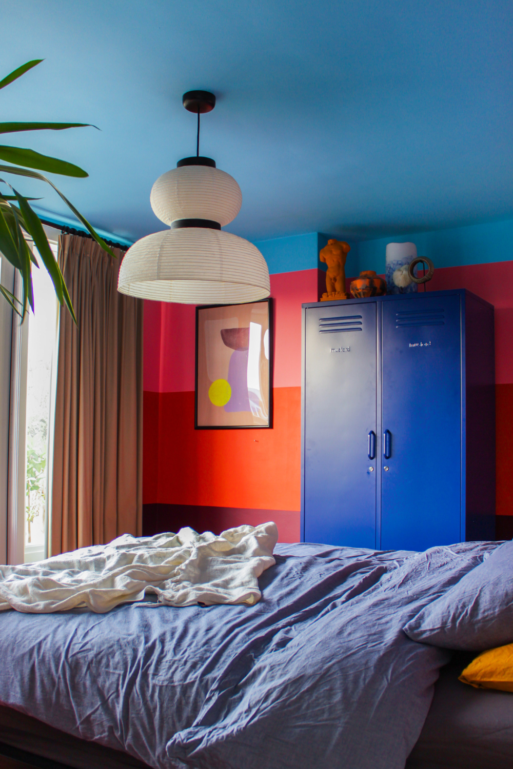 A bedroom is painted in thick horizontal stripes of plum, red and pink, with a light blue ceiling. There is a Navy Twinny locker next to an abstract print.