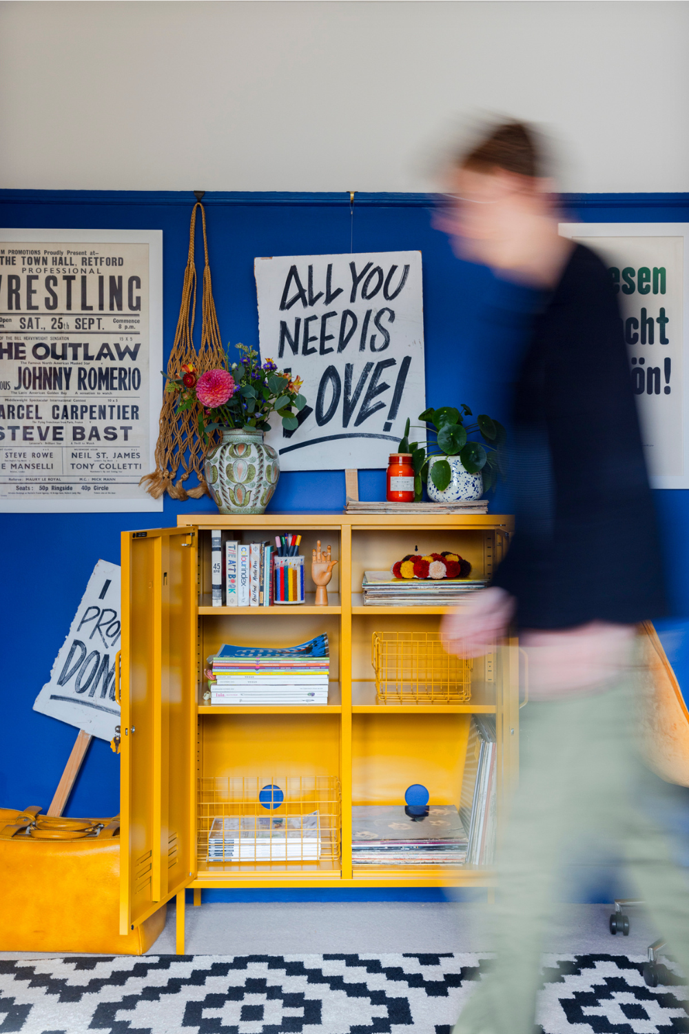 A Mustard Midi locker sits against a cobalt blue wall with bold, black and white graphic prints. One reads 'all you need is love!'