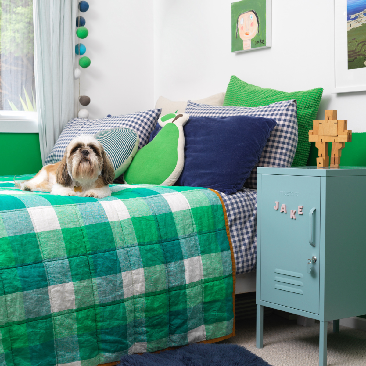 An Ocean Shorty sits next to the bed in a room painted vivid apple green and white. The bedding combines navy gingham with bright green patterns and a whimsical pear cushion, and there is a dog on the bed.