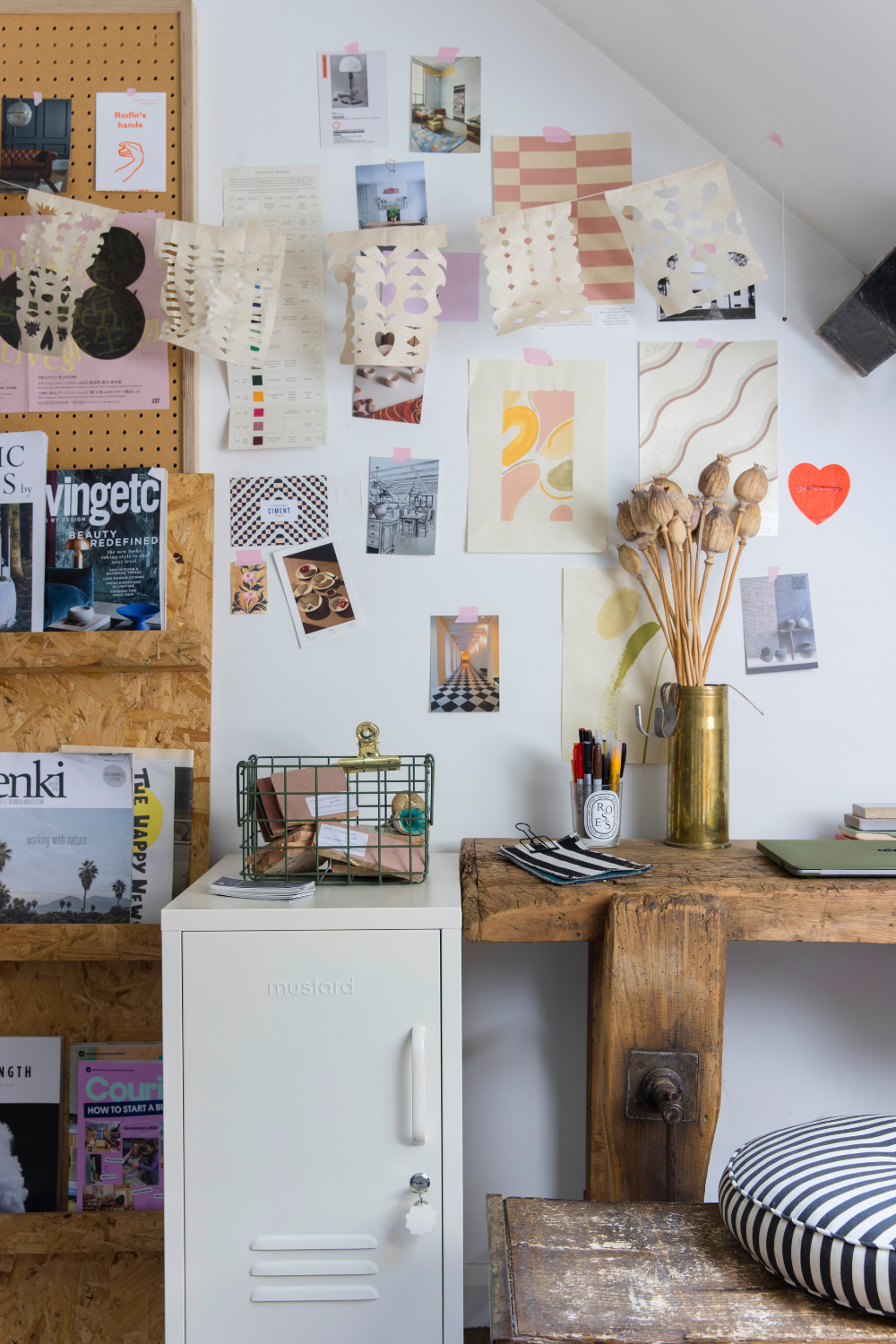 A white wall is covered in colourful photographs and references torn from magazines, scrap-book style. There is a White Shorty next to a wooden desk and a string of papers with lace-like cutouts hung above it.