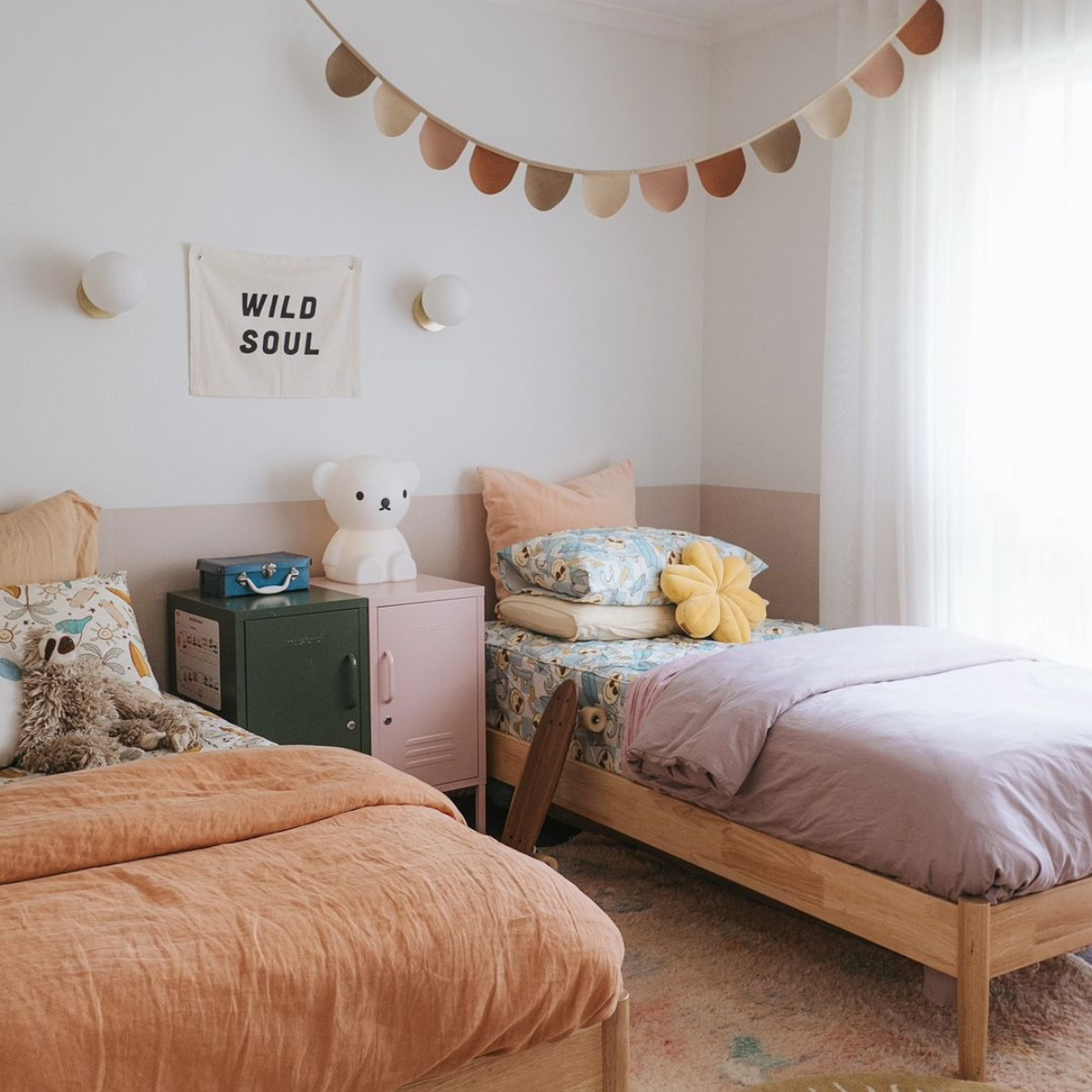 Soft light filters through sheer curtains to a room with two single wooden beds dressed in soft, earthy shades of terracotta and lilac. Between them sits a pairing of Shortys in Olive amd Blush and there is a string of bunting on the ceiling.