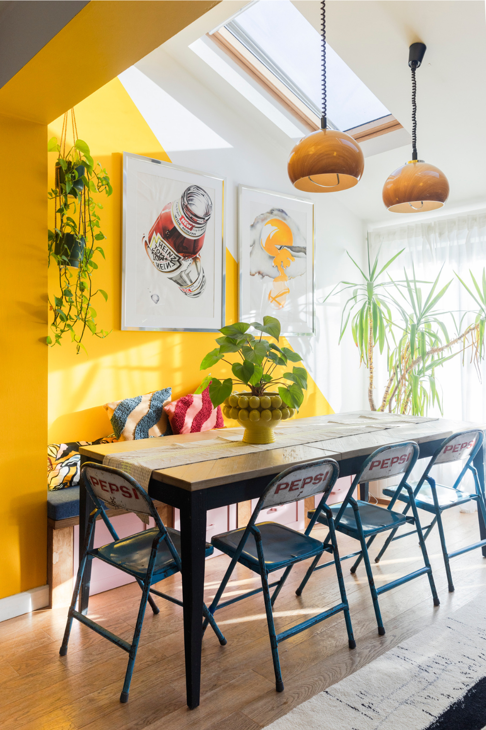 In a yellow dining room there is a large table positioned under a skylight. Vintage metal chairs with the Pepsi logo are tucked around the table.