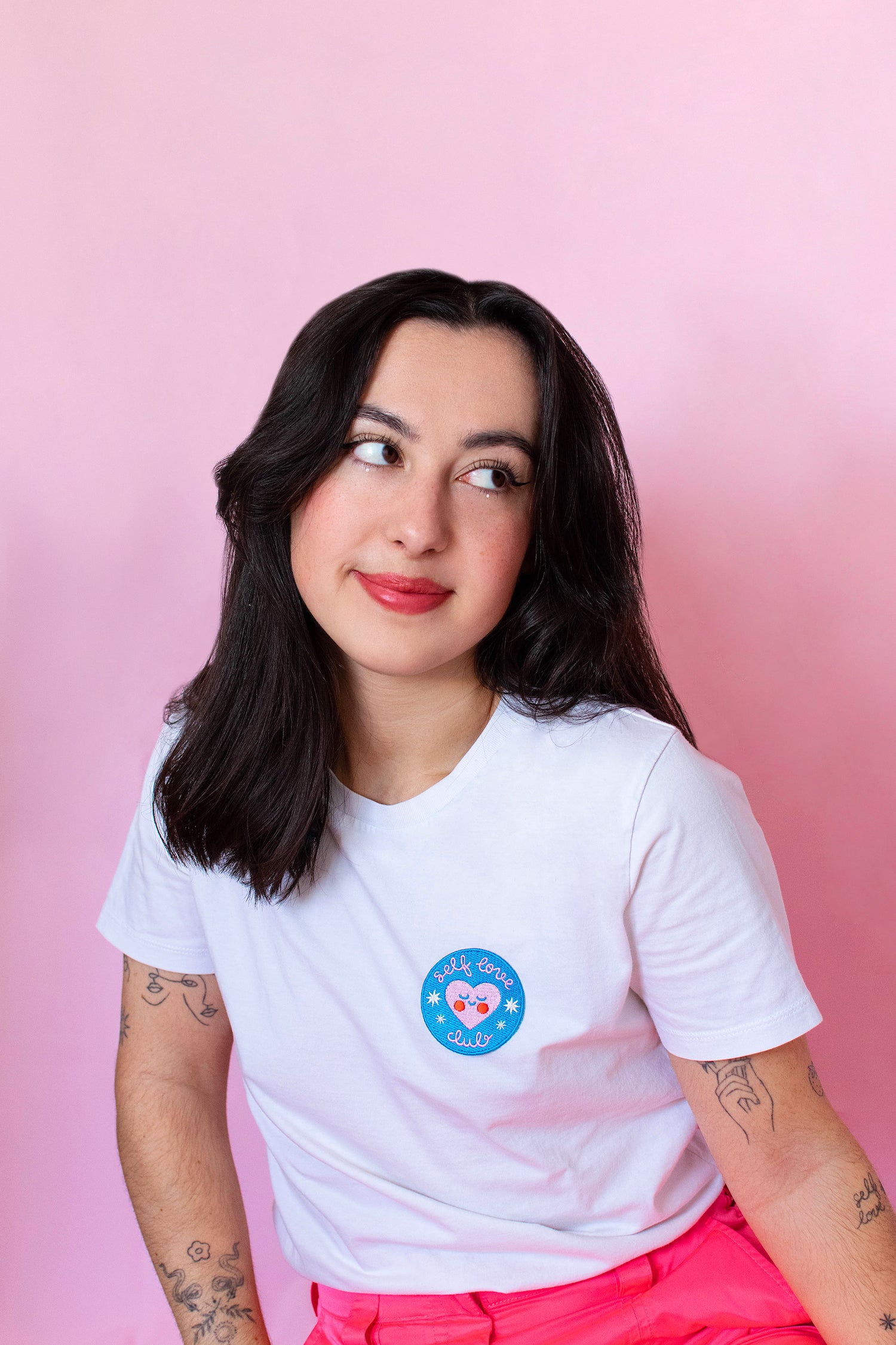 A portrait of the artist. She has dark shoulder length hair and fine line tattoos, and wears a white shirt that reads 'self love club.'