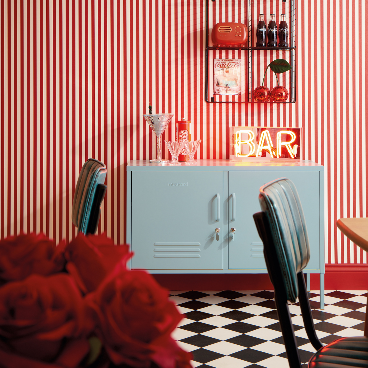 An Ocean Lowdown acts as a bar cabinet in a room with red and white candy-striped wallpaper. There is a red neon bar sign and vintage styling including coke bottles and harlequin flooring.