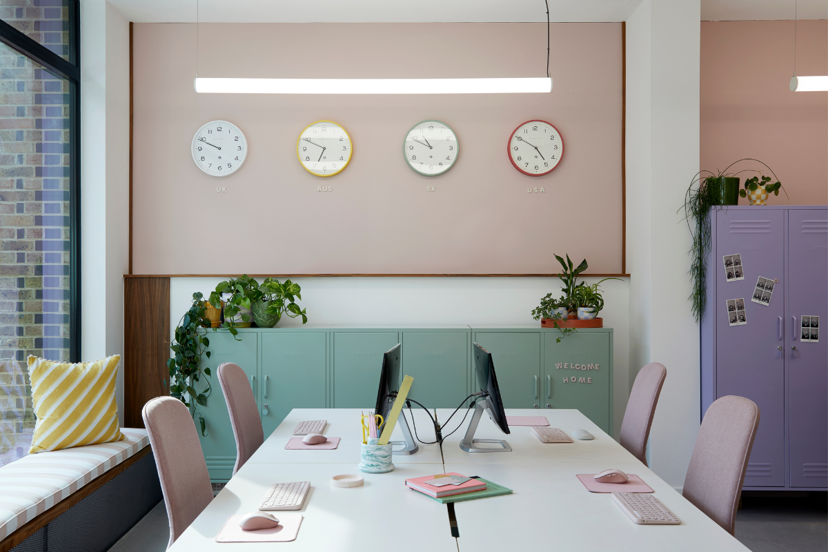 Four colourful clocks hang on a blush pink wall. Beneath it is a large white shared desk space, surrounded by Lilac and Sage lockers.