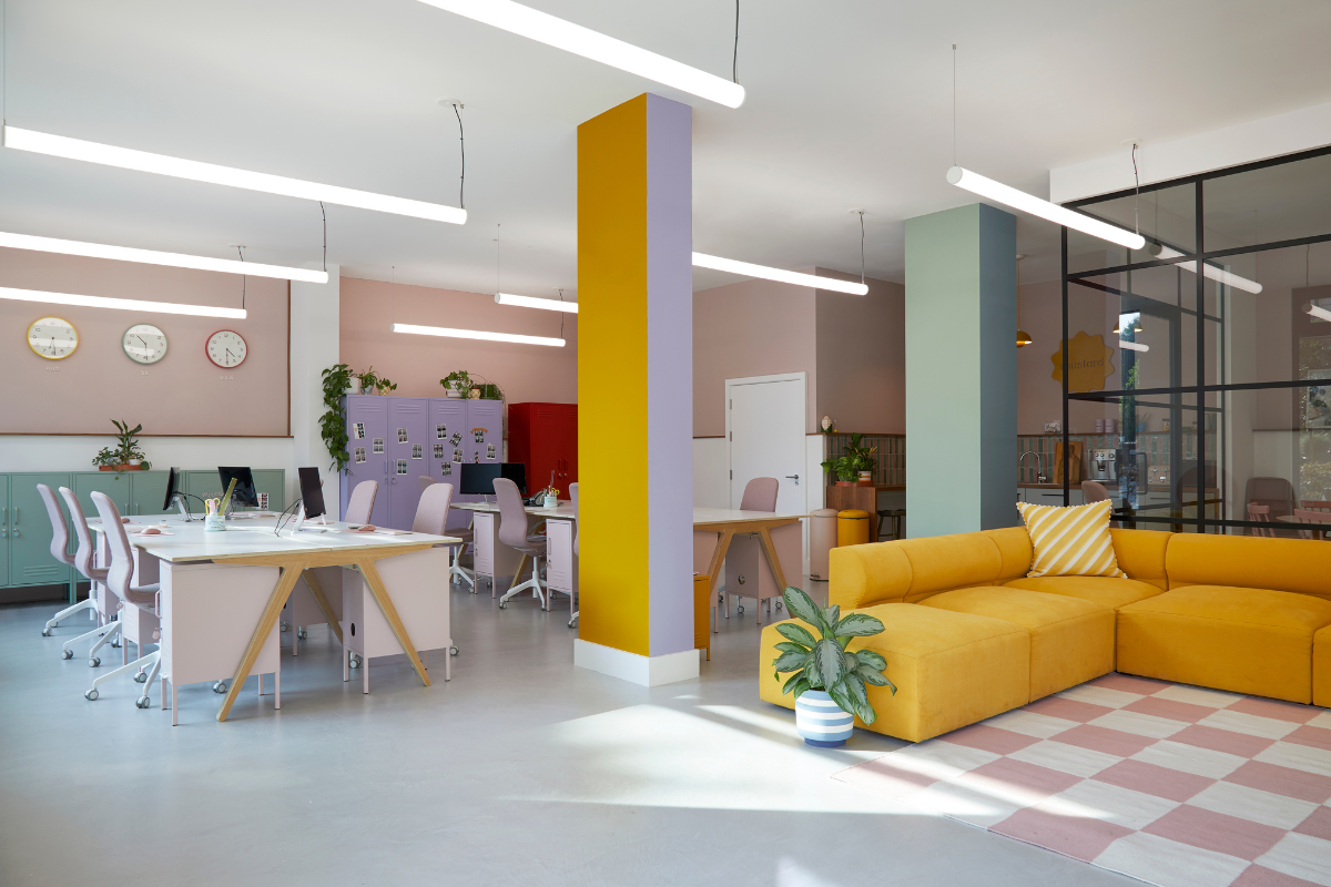 A tall, colourful pillar stands in the centre of the space. One side is painted Mustard, the other Lilac. To the right there is a large, L shaped mustard sofa and a white and pink checkerboard rug.