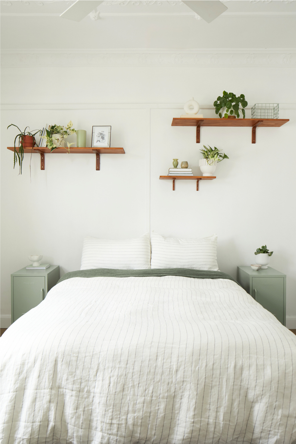 In a white bedroom, two Sage Shortys sit next to a bed dressed in pinstripe linen. There are three wooden floating shelves on the wall with trailing plants and books arranged artfully.