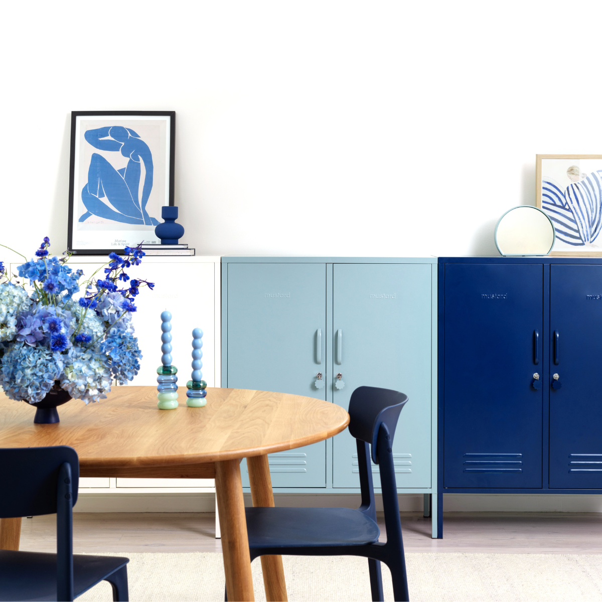 A trio of Midi lockers in an ombre pattern from white to Ocean to Navy are styled behind a round wooden dining table. There is a vase of luscious, full blue flowers and a selection of blue-toned artworks.