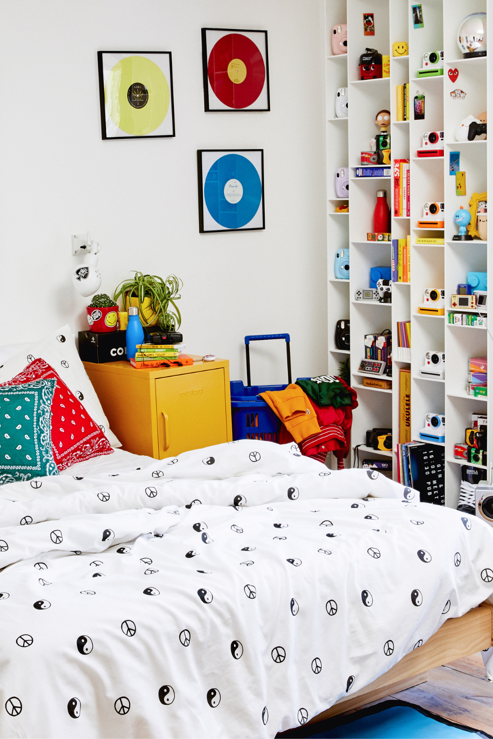 A Mustard Shorty sits between a bed and a bookshelf covered in brightly colored polaroid cameras. There are colorful records framed on the wall.