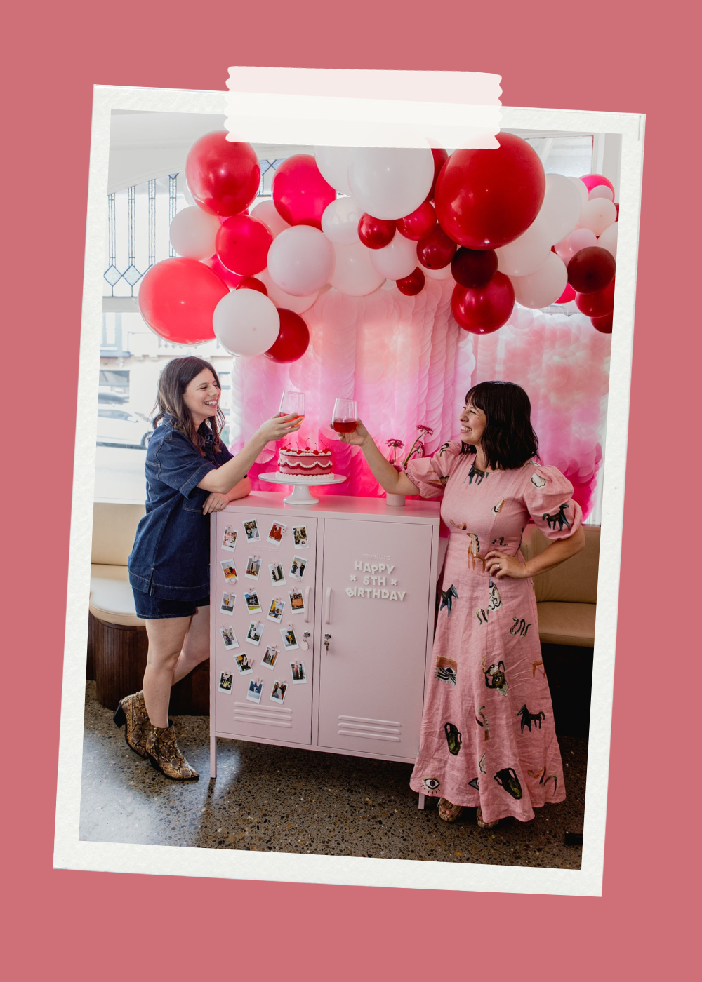 Becca and Jess clink glasses under a balloon garland. They are leaning on a Blush locker which has a cake on it.