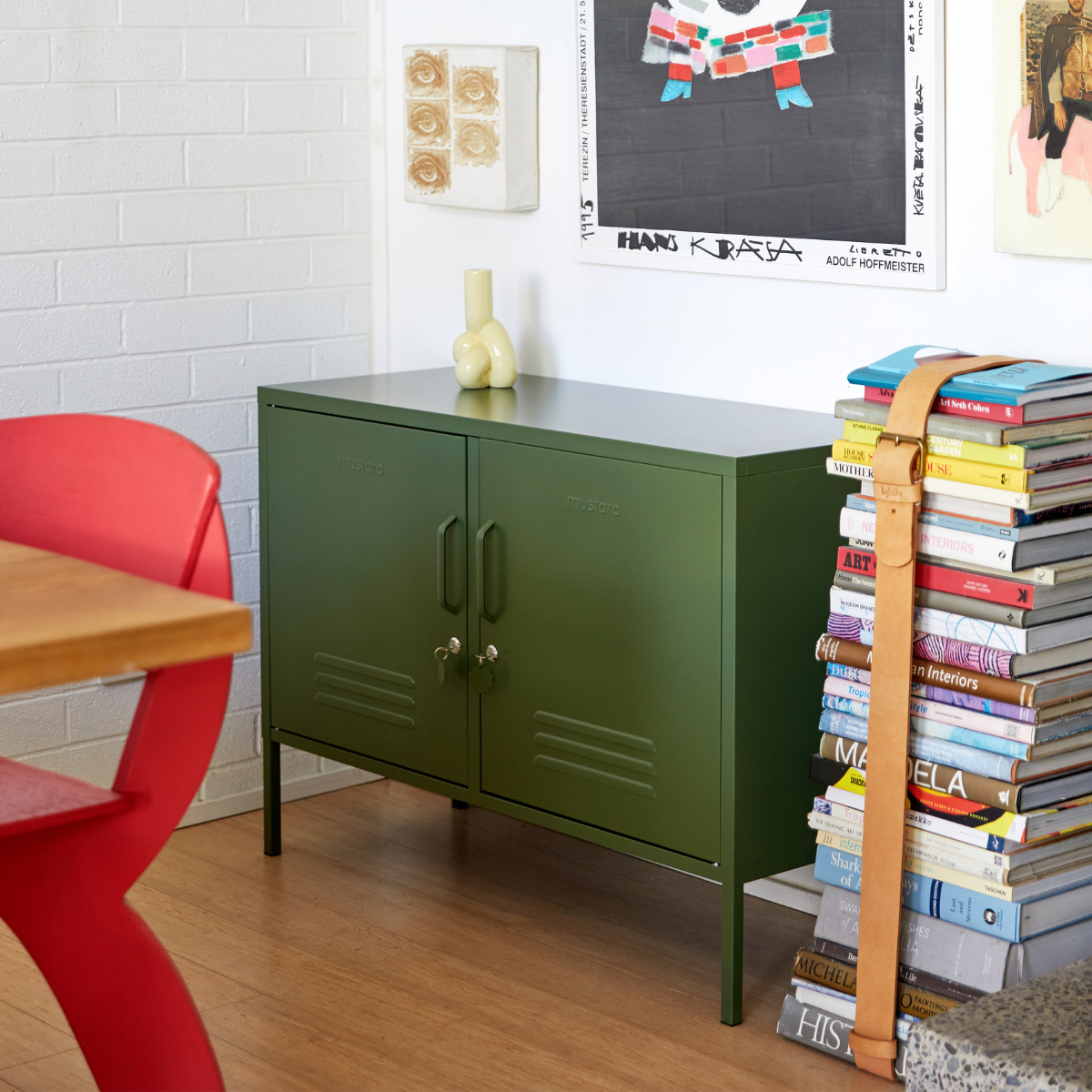 An Olive Lowdown sits next to a stack of colourful hardback books bound with a leather strap. There is bright artwork on the walls and a feature red chair.