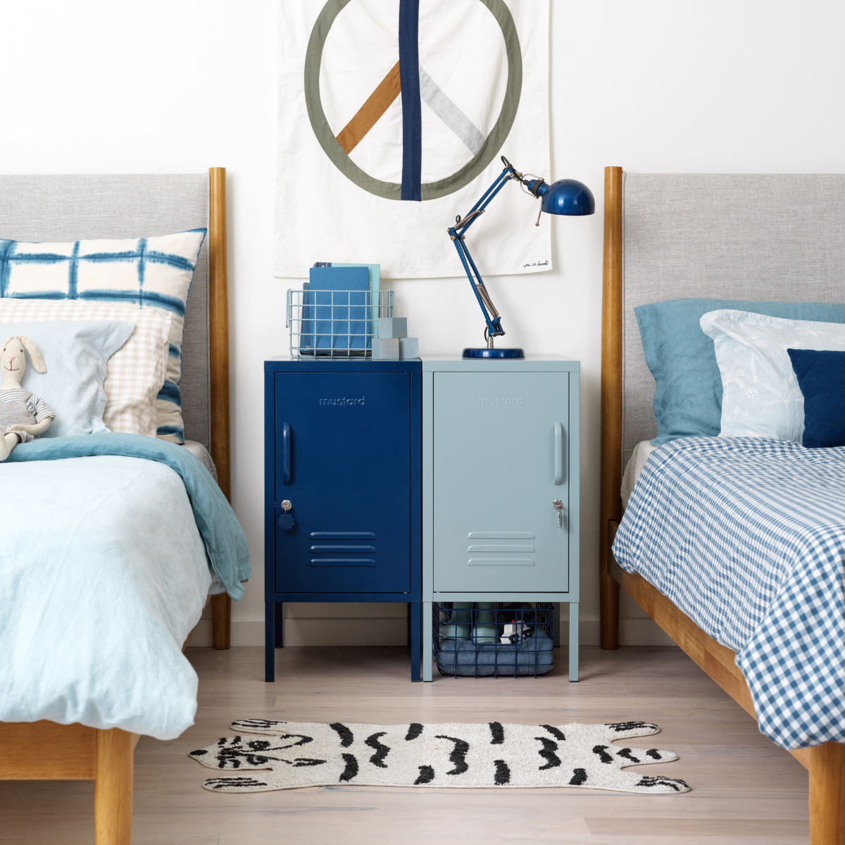 Two single beds sit either side of a pair of blue Shortys side by side. One locker is Ocean and one is Navy. The beds are styled with patterned fabrics in blues and whites and there is a small black and white rug on the floor.