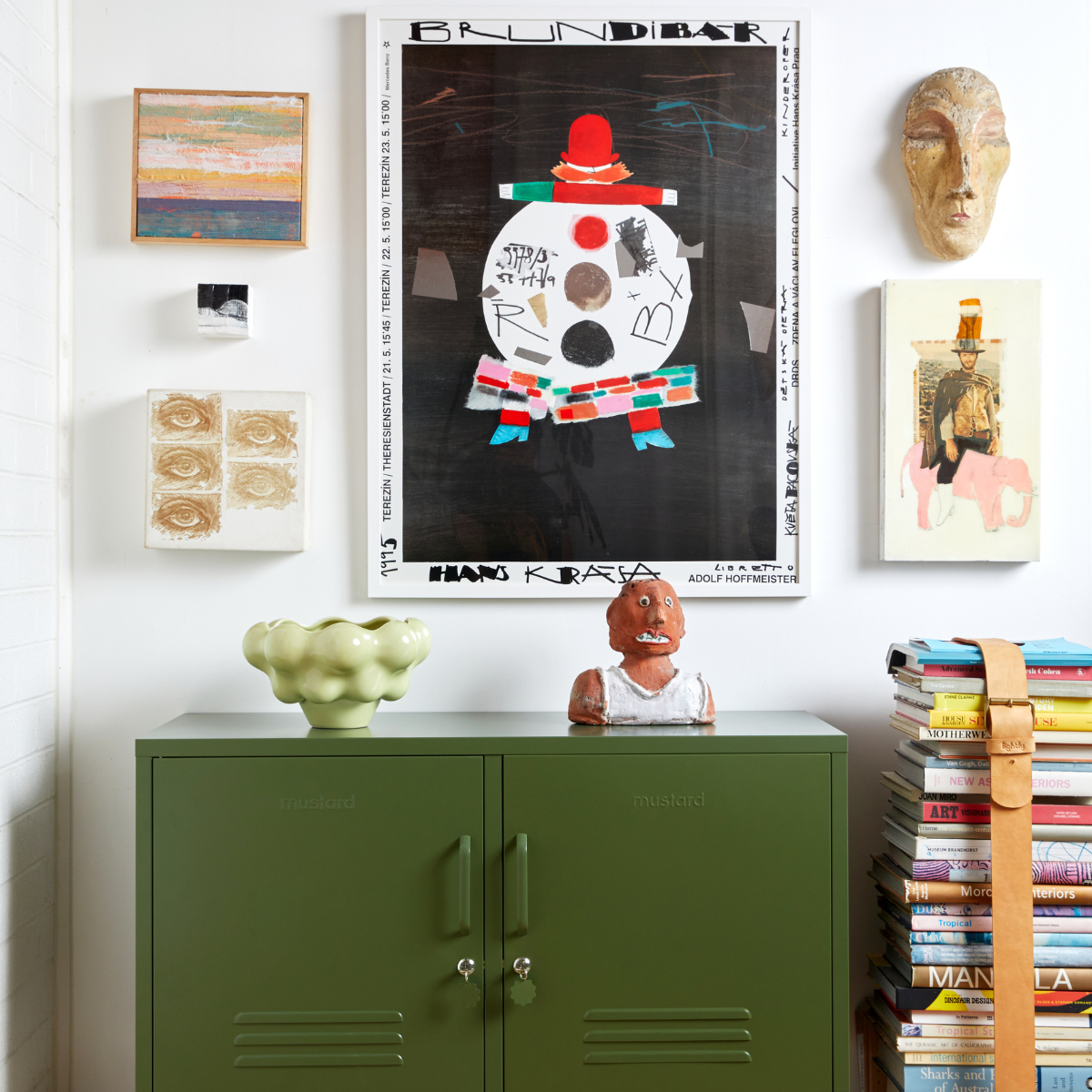 An olive green locker houses some ceramics and sits next to a tall stack of books bound by a leather belt. Bright artworks are featured on the wall.