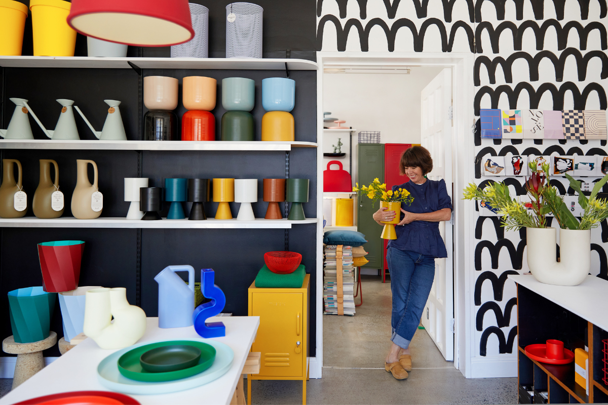 Nina holds a vase of yellow flowers as she leans against a door frame inside MiiO Store. Colourful objects surround her on the shelves.