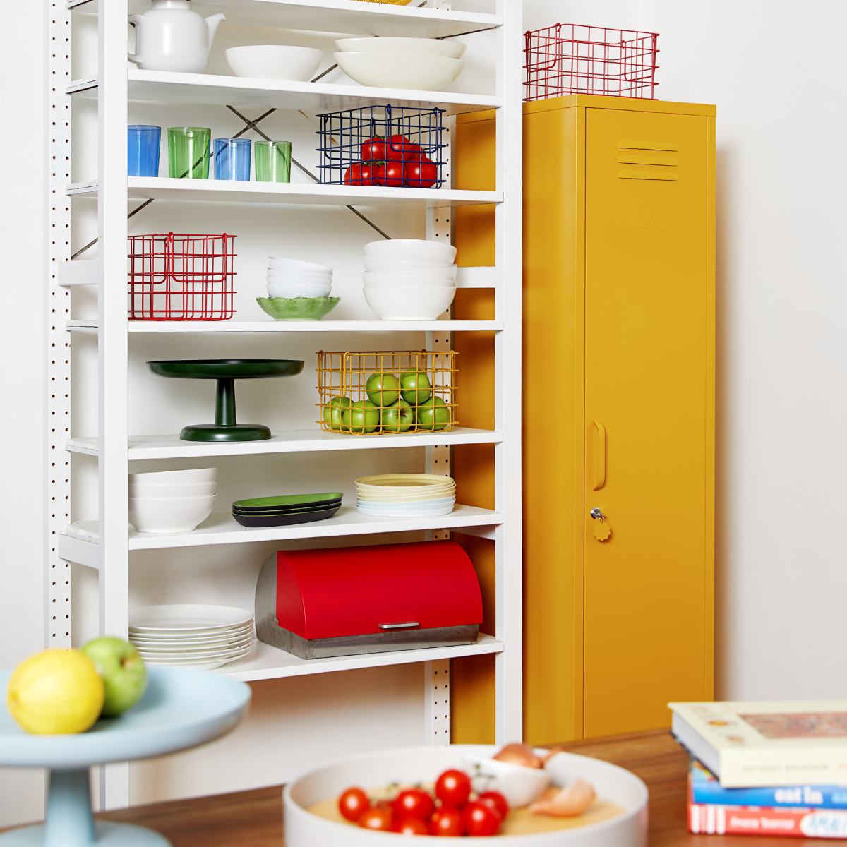 A set of open shelving in the kitchen displays crockery and glassware in primary colours. There is a mustard yellow locker in the corner and brightly coloured baskets display fruit and vegetables arranged by colour.