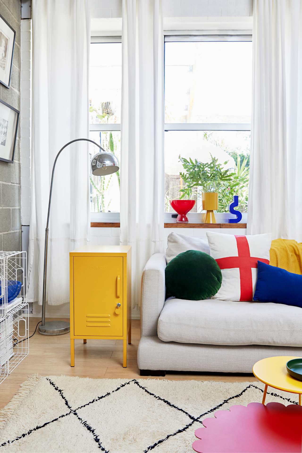 A light grey couch is scattered with cushions in primary colours and a silver floor lamp curves over a mustard yellow locker. Through the window is a view of trailing greenery.