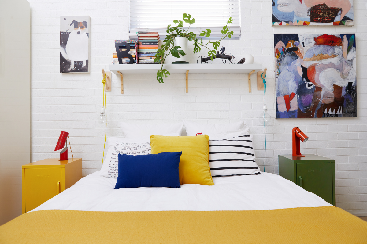 A bed is dressed in white linens with mustard yellow, blue and striped accents. A small locker with a little red lamp sits on each side of the bed and above it is a shelf featuring stacks of books and a trailing vine.